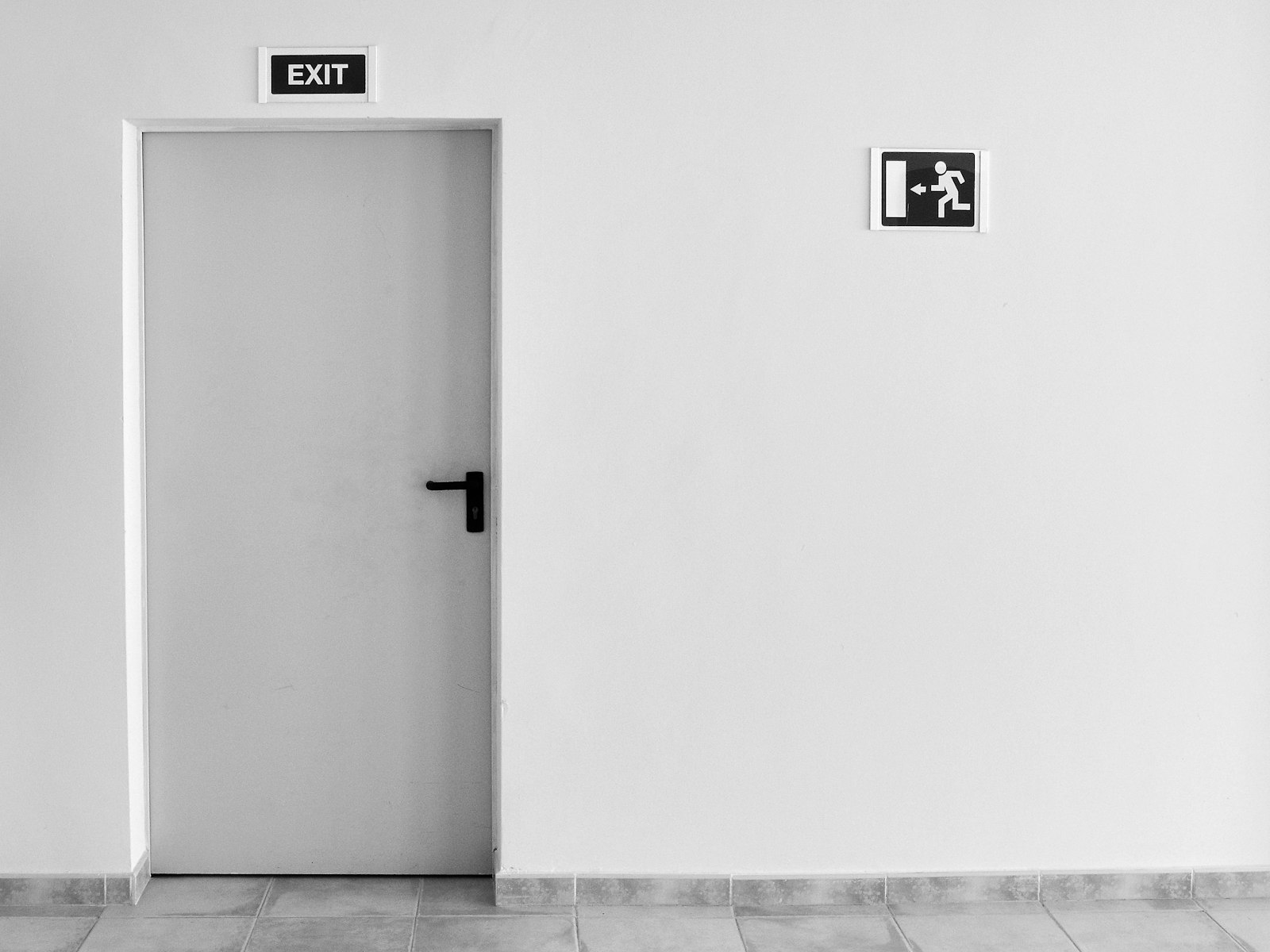 Grey fire door with running-man exit pictogram beside a large white wall